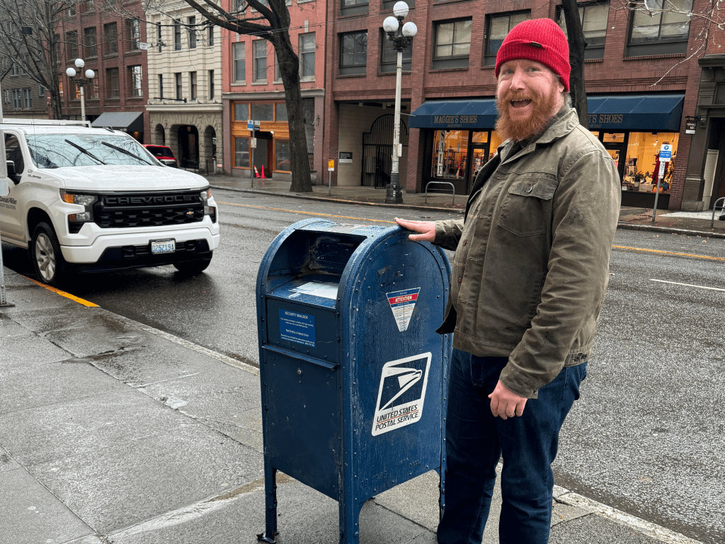 A picture of a white man with a red beard and a red beanie standing next to a mailbox